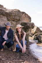 Woman wearing a beige Stevie Wool Wide-Brim Hat  and decorative band, sitting on rocks by the water-Fleastyle.