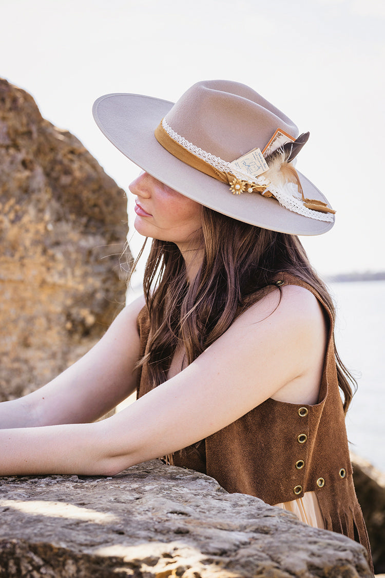 Woman wearing a beige Stevie Wool Wide-Brim Hat  and decorative band, sitting on rocks by the water-Fleastyle.